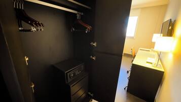 A hotel room with a dark-colored wardrobe, a small desk with drawers, and a bedside table with a lamp.