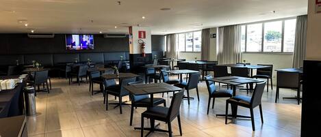 A spacious restaurant with multiple tables and chairs, a flat-screen TV mounted on the wall, and large windows allowing natural light.