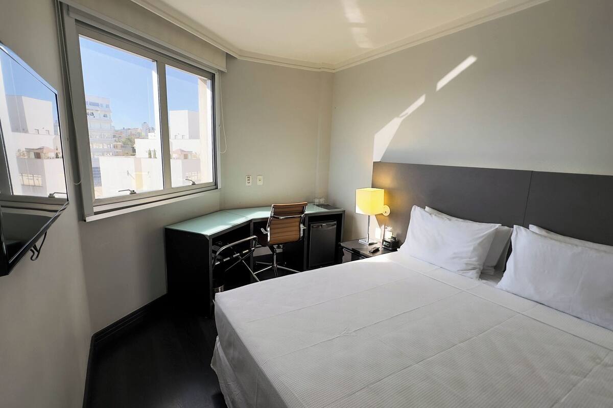 A hotel room with a bed, a desk, a chair, a desk lamp, and a window with a view of buildings.