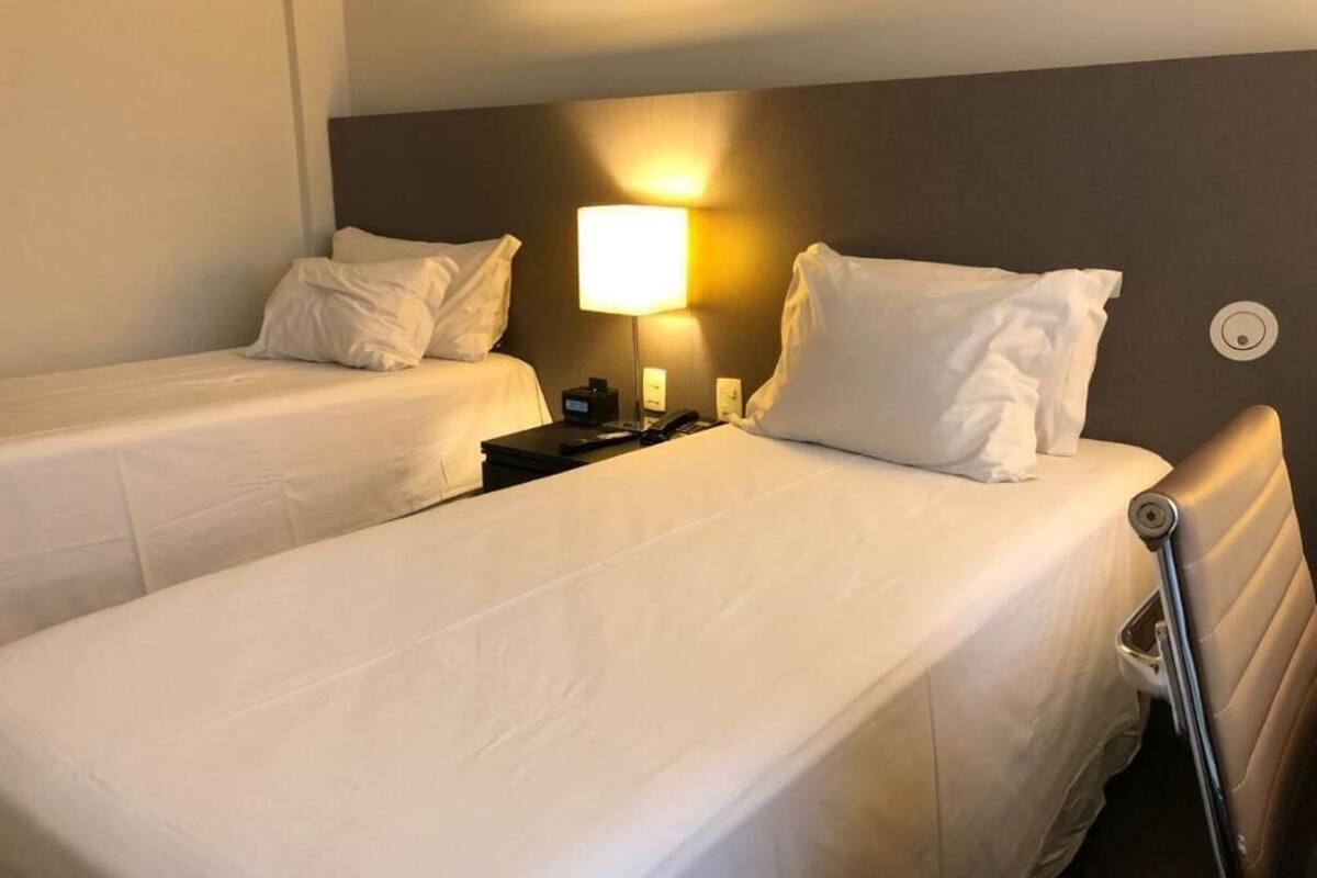 A hotel room with two single beds, a nightstand, a lamp, and a wall-mounted radiator.