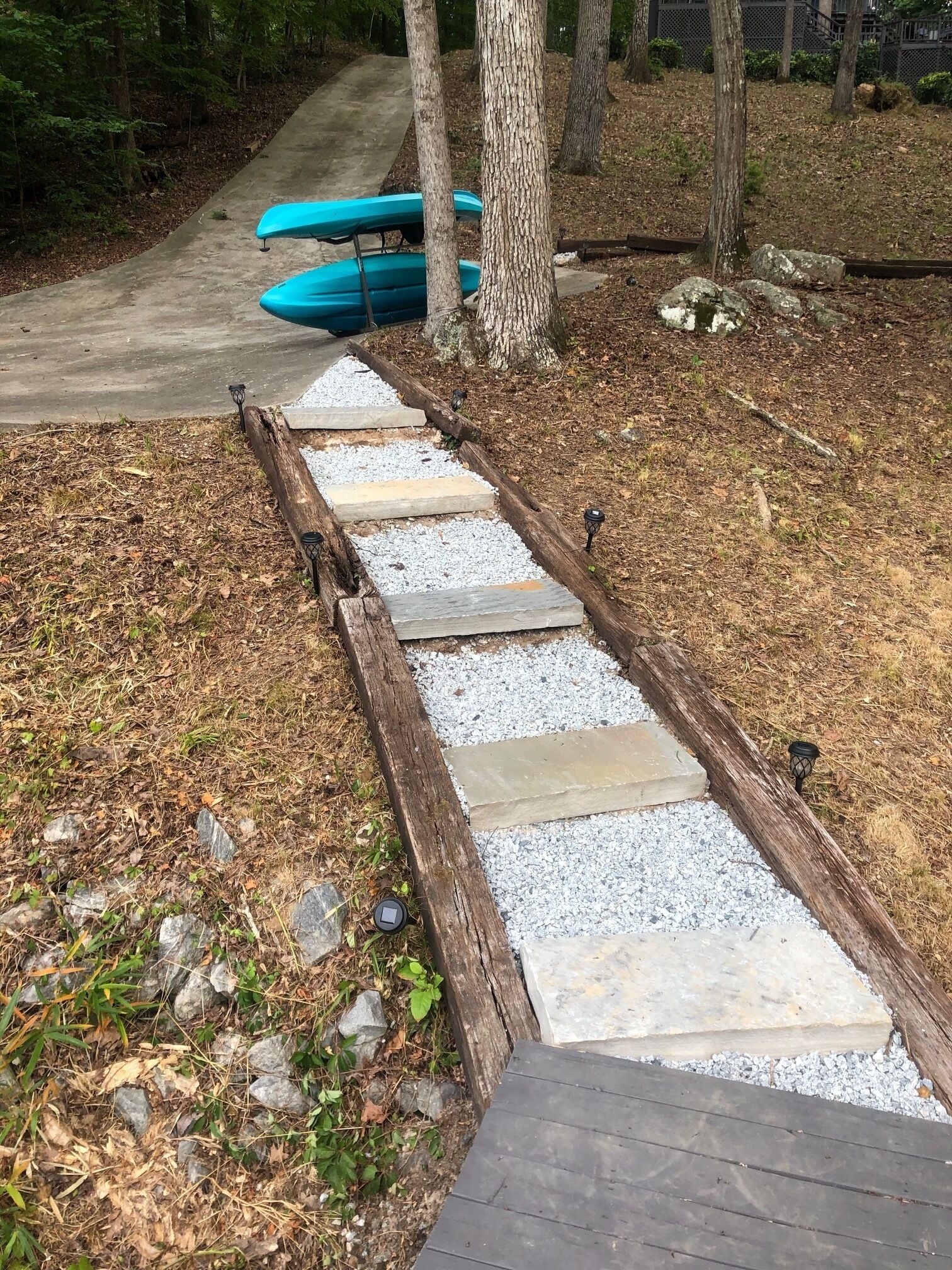 Ein Weg mit Holstufen und Kies, der zu einem Betonpfad führt, im Hintergrund sind Kajaks zu sehen.