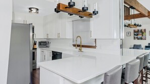 Una cocina moderna con armarios blancos, una isla grande con un lavamanos, horno empotrado y microondas. El techo tiene vigas de madera e iluminación colgante sobre la isla.