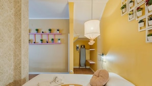 A hotel room with a bed, a bedside table with a tray, a hanging light fixture, a shelf with plants, and a door leading to a bathroom. - Y.Quebedo - Boutique Apartments - Poetico (Setúbal)