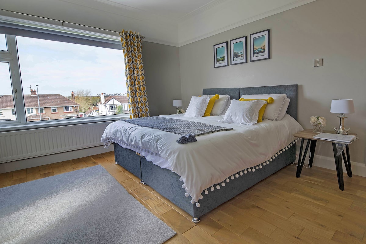 A bedroom with a large bed, a window with curtains, a radiator, and a view of houses outside.
