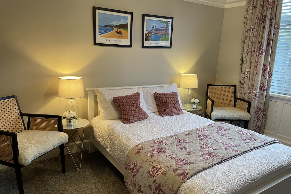 A neatly arranged hotel room with a large bed, two side chairs, a small table with a lamp, and two framed pictures on the wall.
