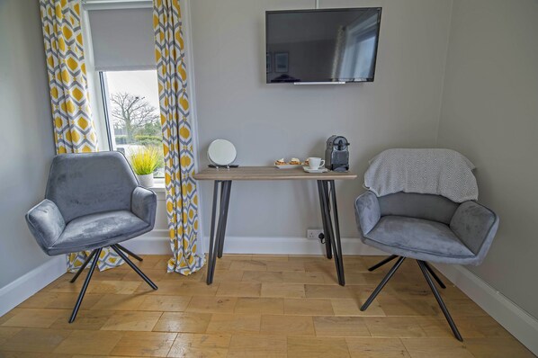 Ein Zimmer mit zwei grauen Stühlen, einem kleinen Tisch mit Tasse und Gebäck, einem an der Wand montierten Fernseher und einem Fenster mit gemusterten Vorhängen.