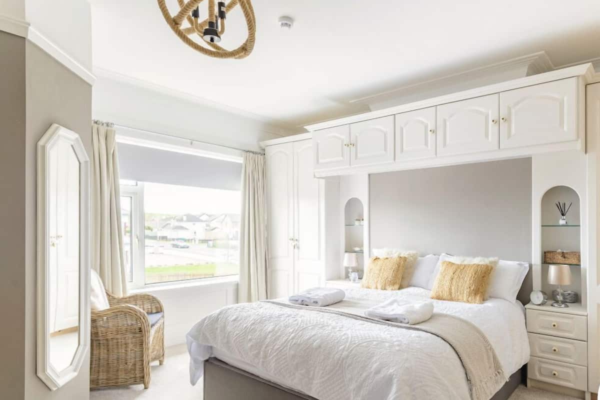 A bedroom with a large bed, a chest of drawers, a wicker chair, and a view of the outside.
