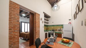 Cocina compacta con pared de ladrillo, zona de comedor con una computadora portátil y frutero, y un refrigerador a la vista.