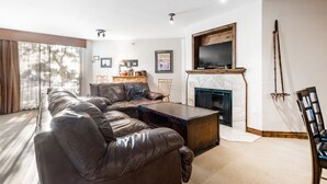 Una sala de estar con sofá de cuero, mesa de centro de madera, chimenea y televisor montado en la pared.