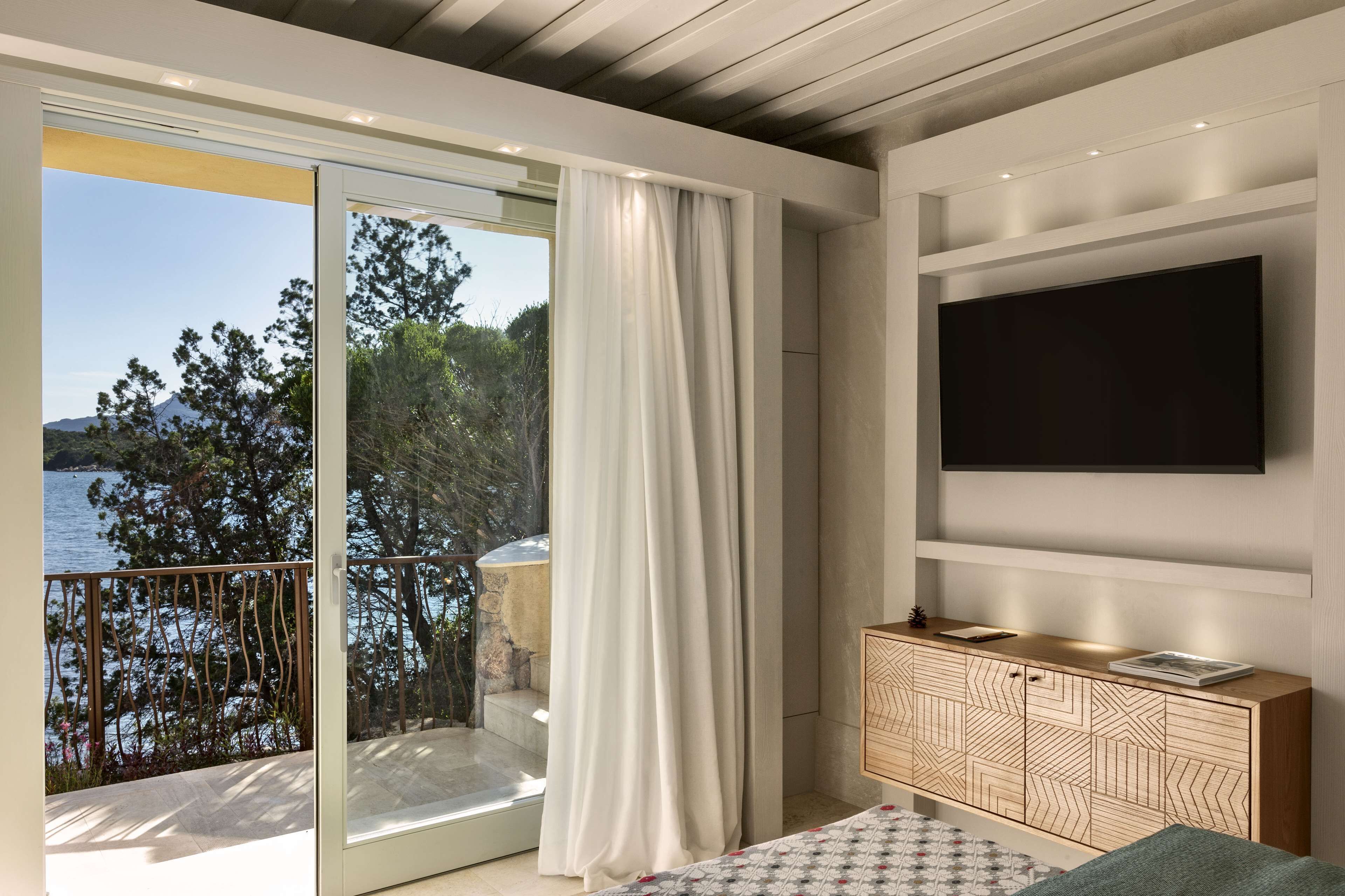 Ein modernes Zimmer mit einer großen Glasschiebetür, die auf einen Balkon mit Blick auf das Meer und Bäume führt. An der Wand ist ein Flachbildfernseher angebracht und es gibt ein Holzschränkchen mit Schubladen.