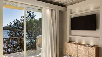 Ein modernes Zimmer mit einer großen Glasschiebetür, die auf einen Balkon mit Blick auf das Meer und Bäume führt. An der Wand ist ein Flachbildfernseher angebracht und es gibt ein Holzschränkchen mit Schubladen.