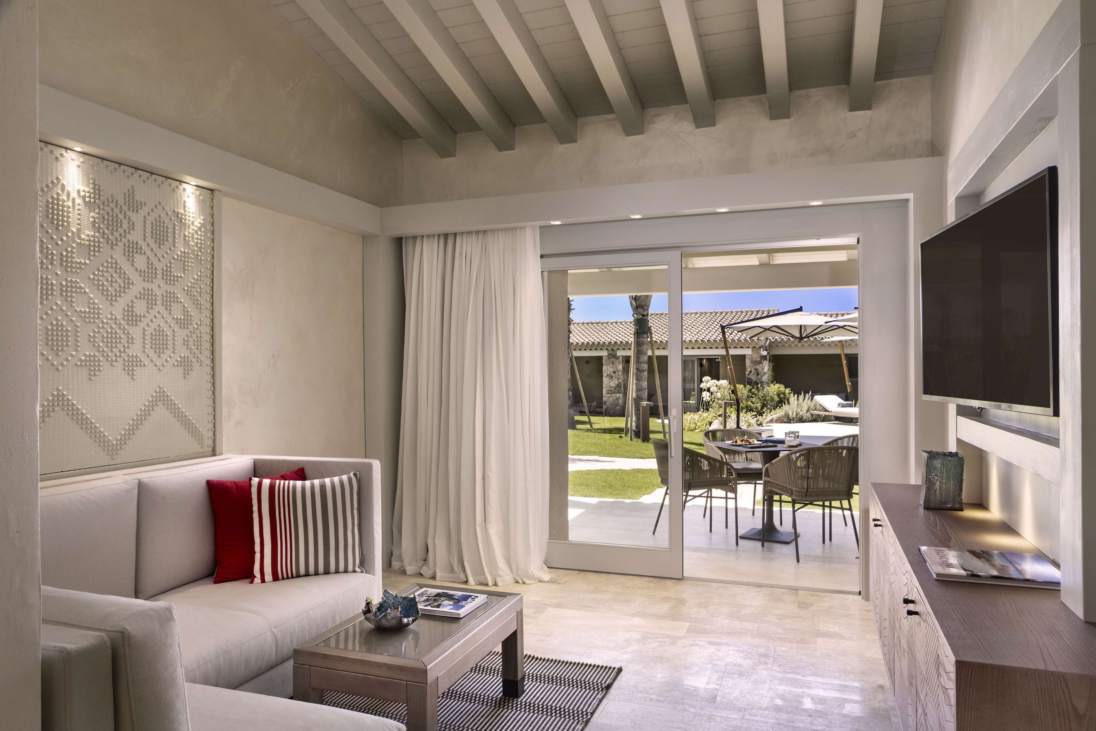 Una sala de estar moderna con un sofá, una mesa de centro y un televisor. Hay un ventanal corredero que da a un patio al aire libre con una mesa y sillas de comedor.