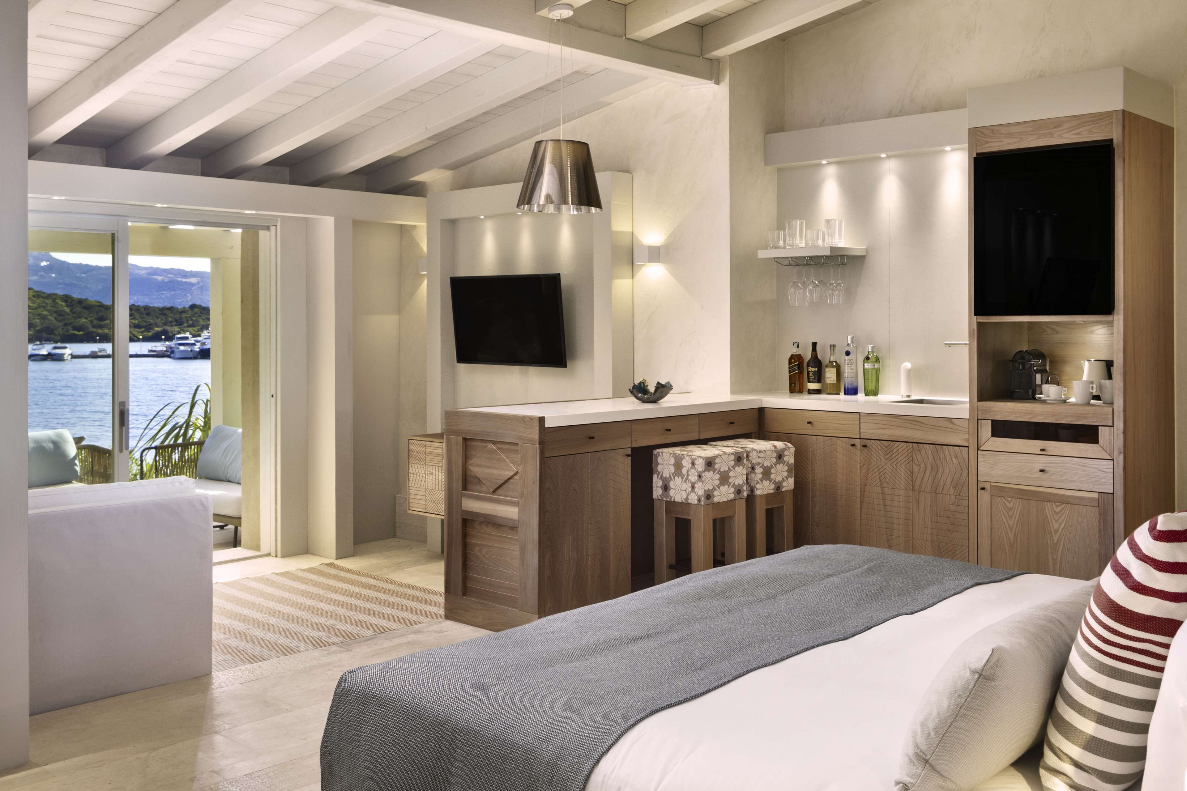 Ein modernes Schlafzimmer mit einem großen Bett, einem an der Wand montierten Fernseher, einem Holztisch mit Stuhl und Meerblick durch eine Schiebetür.