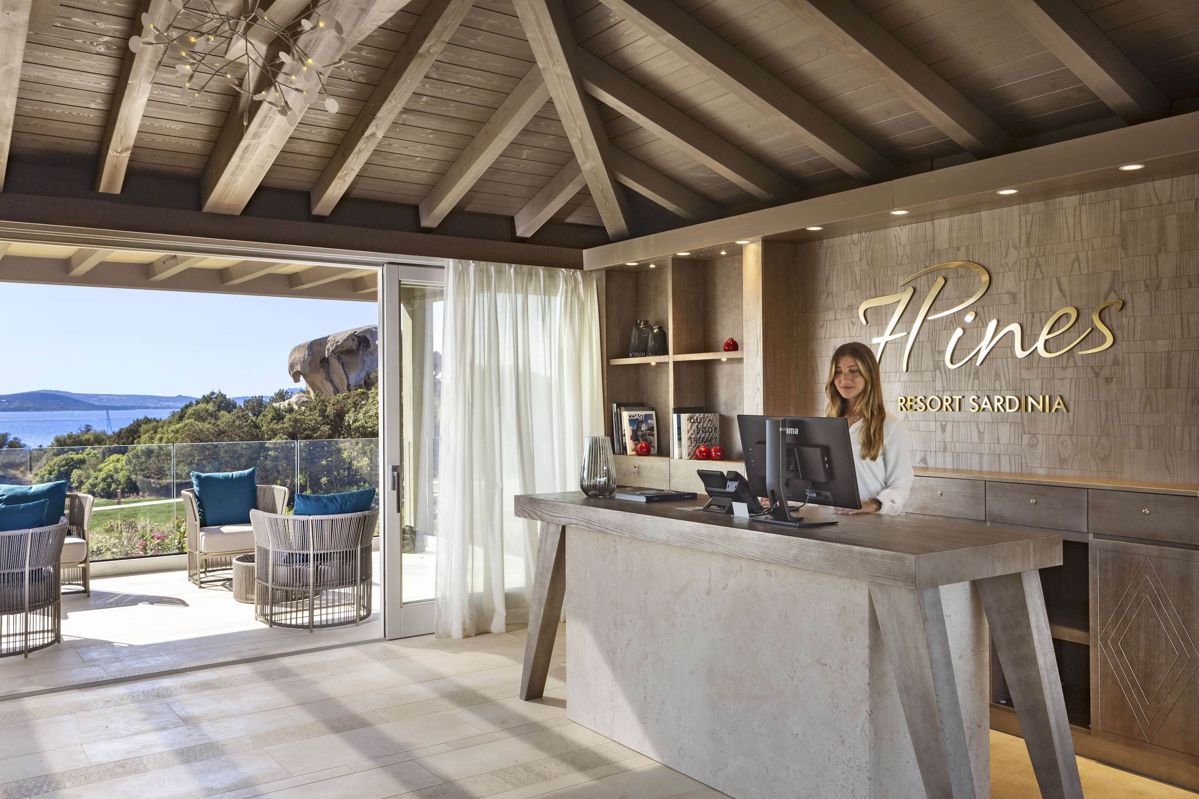 Eine Rezeption mit Blick auf das Meer und die Berge, ein modernes Interieur mit Holzbalkendecke und einem Schild mit der Aufschrift 'Hines Resort Sardinia'.