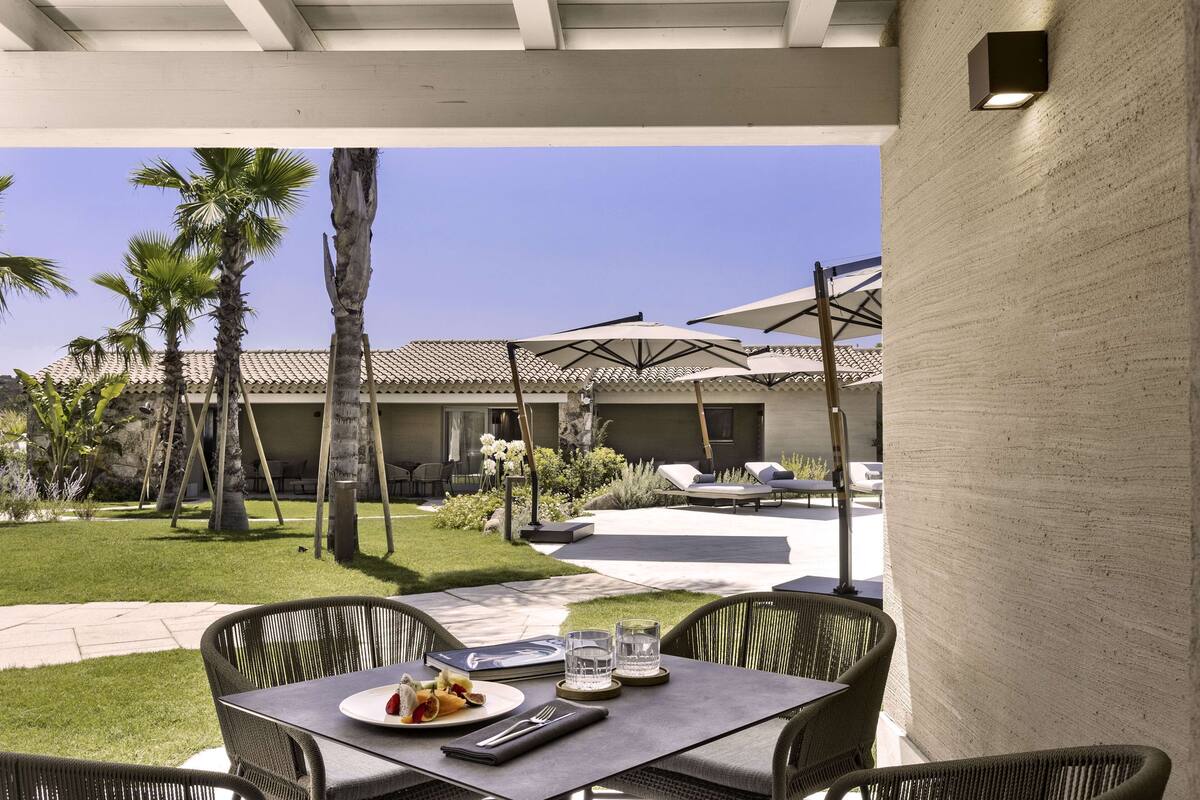 Une terrasse avec une table dressée pour deux personnes, entourée de chaises en osier, donnant sur un jardin avec des palmiers et un bâtiment doté d’un toit en tuiles.