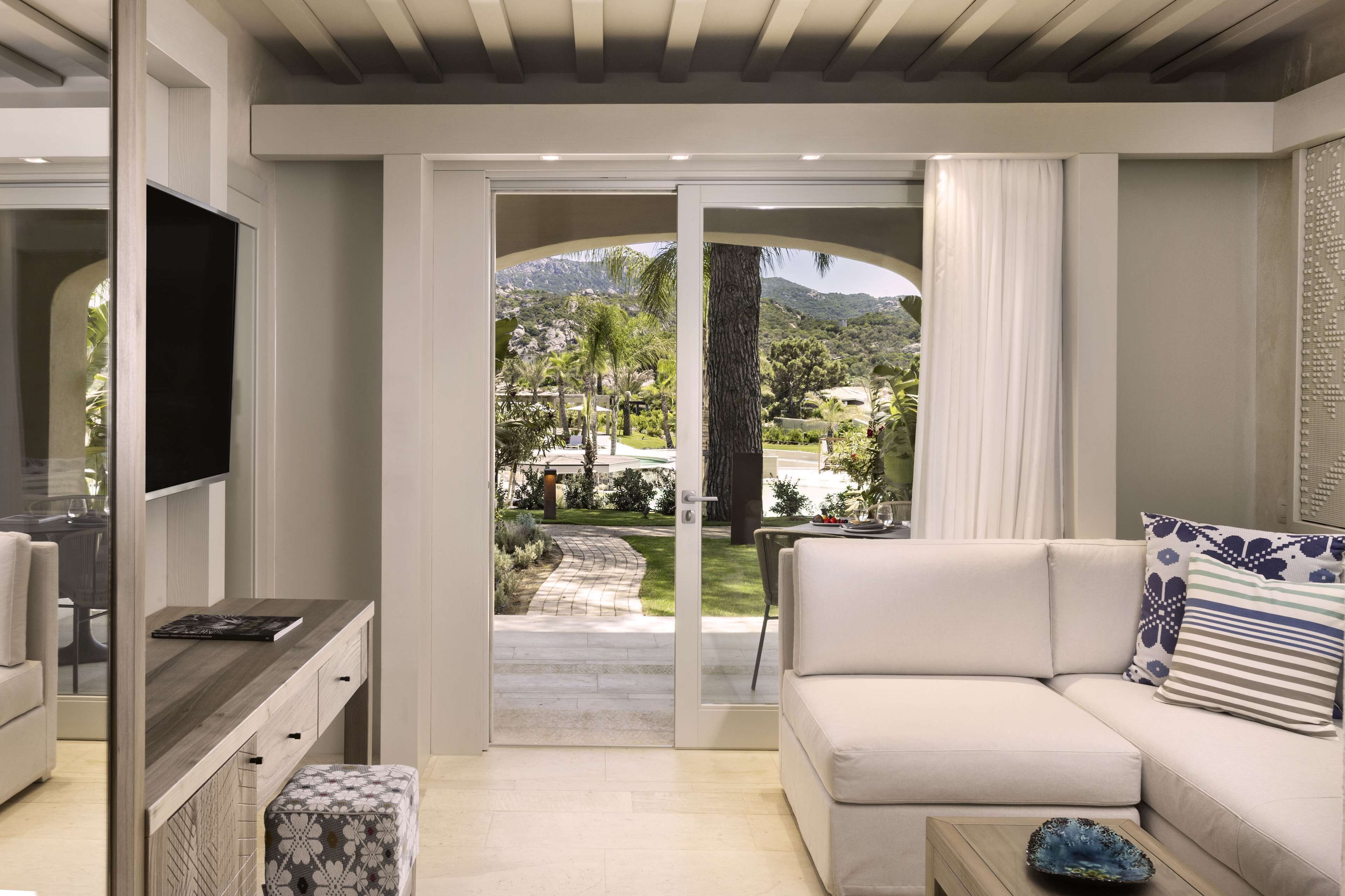 A modern living room with a sofa, a coffee table, and a TV. There is a sliding glass door leading to a garden with a view of mountains.