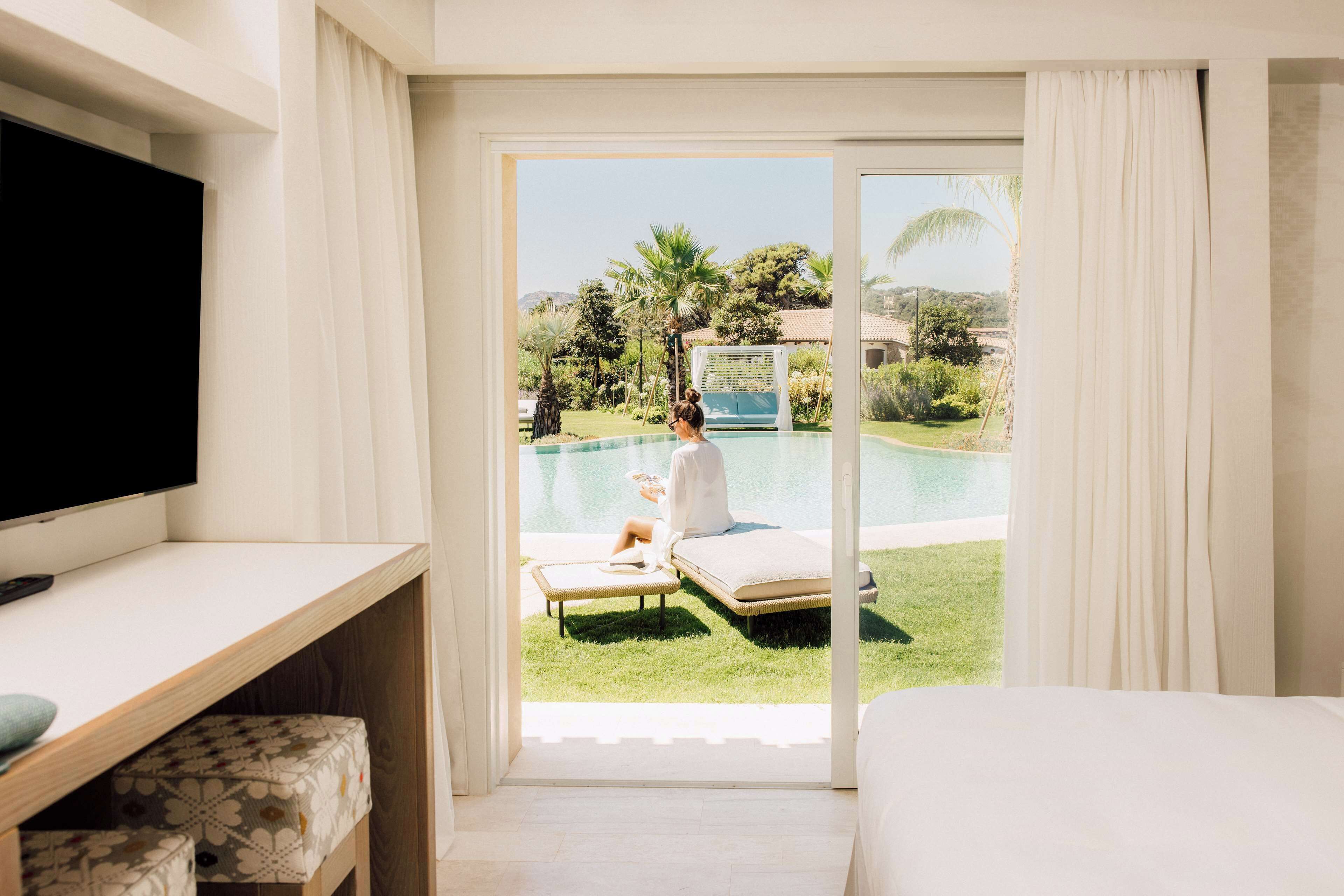 A room with a TV, a bed, a desk, and a view of a pool area with a person relaxing.