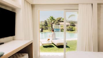 A room with a TV, a bed, a desk, and a view of a pool area with a person relaxing.