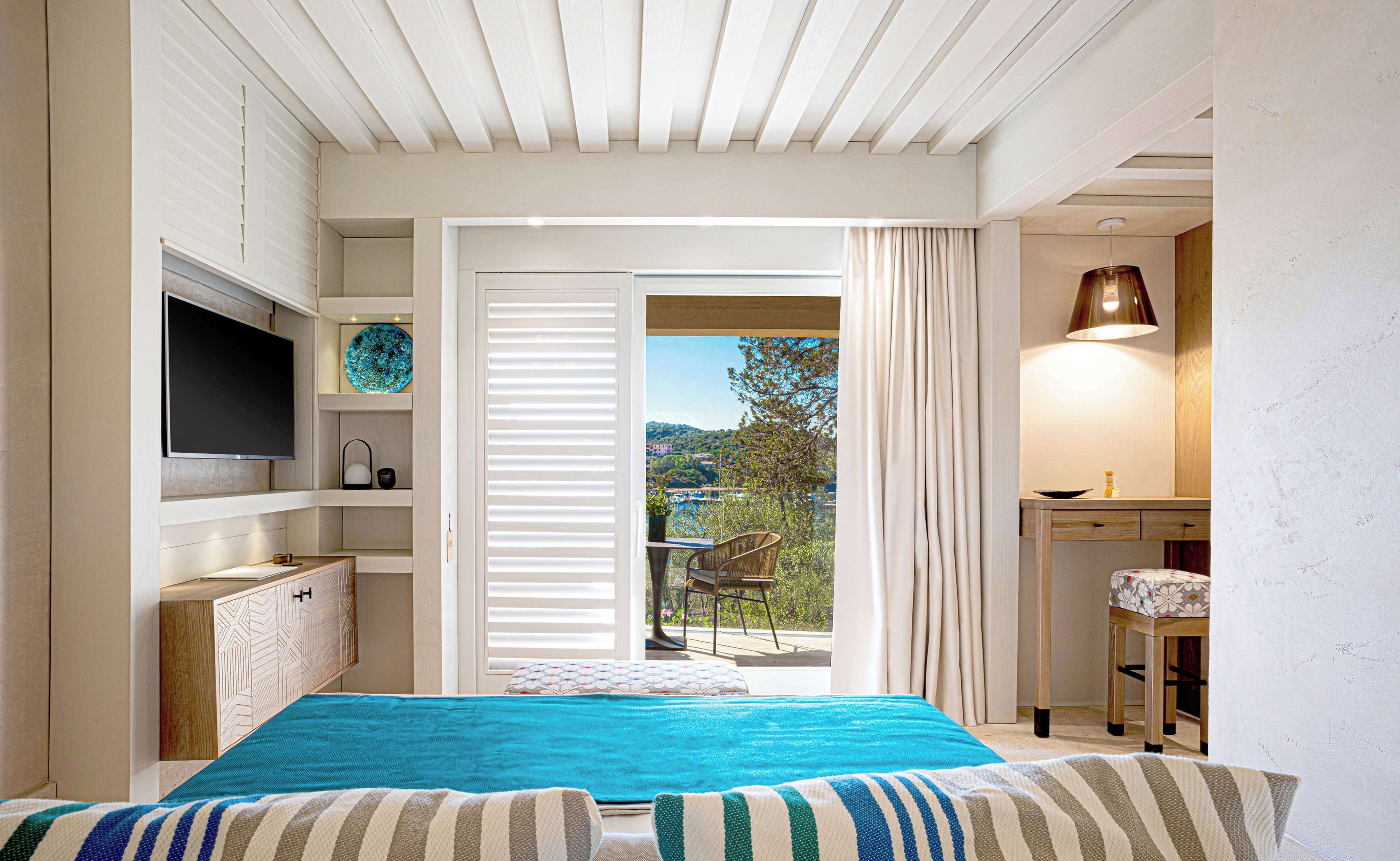 A bedroom with a bed, a TV, a desk, and a view of the outdoors.