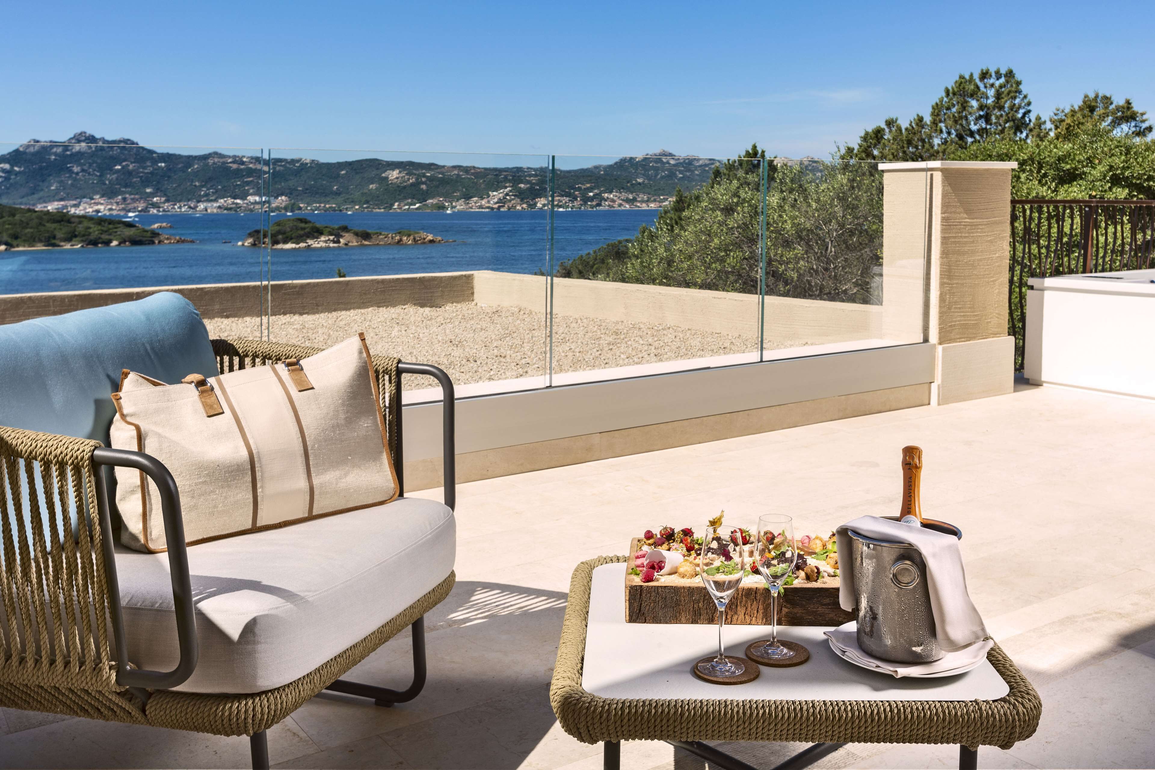 A balcony with a wicker sofa and table, a toaster, and a glass of wine, overlooking a coastal landscape.