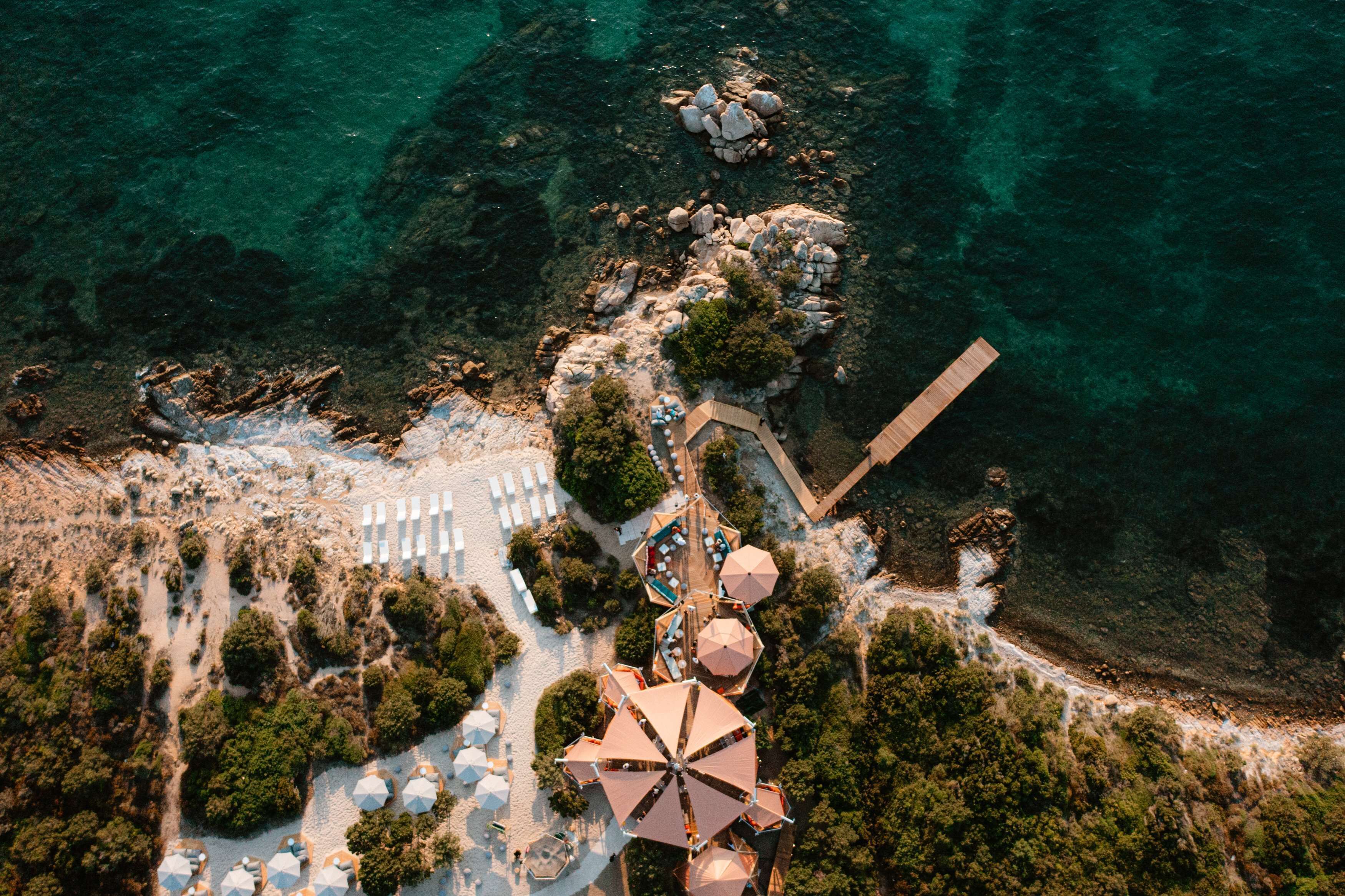 An aerial view of a coastal resort with a beach, umbrellas, and a wooden walkway.