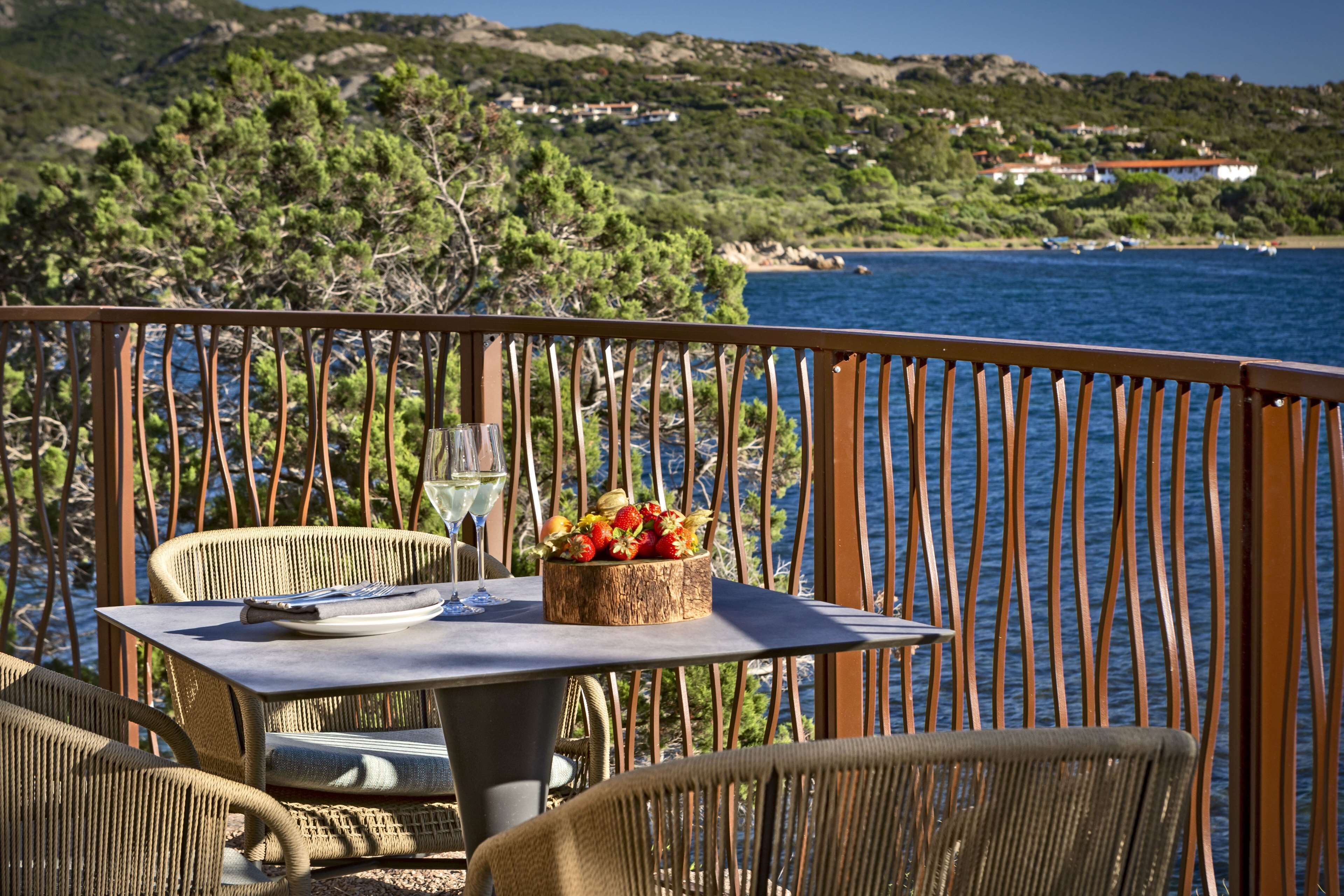 A dining area with wicker furniture, a table with a fruit centerpiece, and a view of the sea and hills in the background.