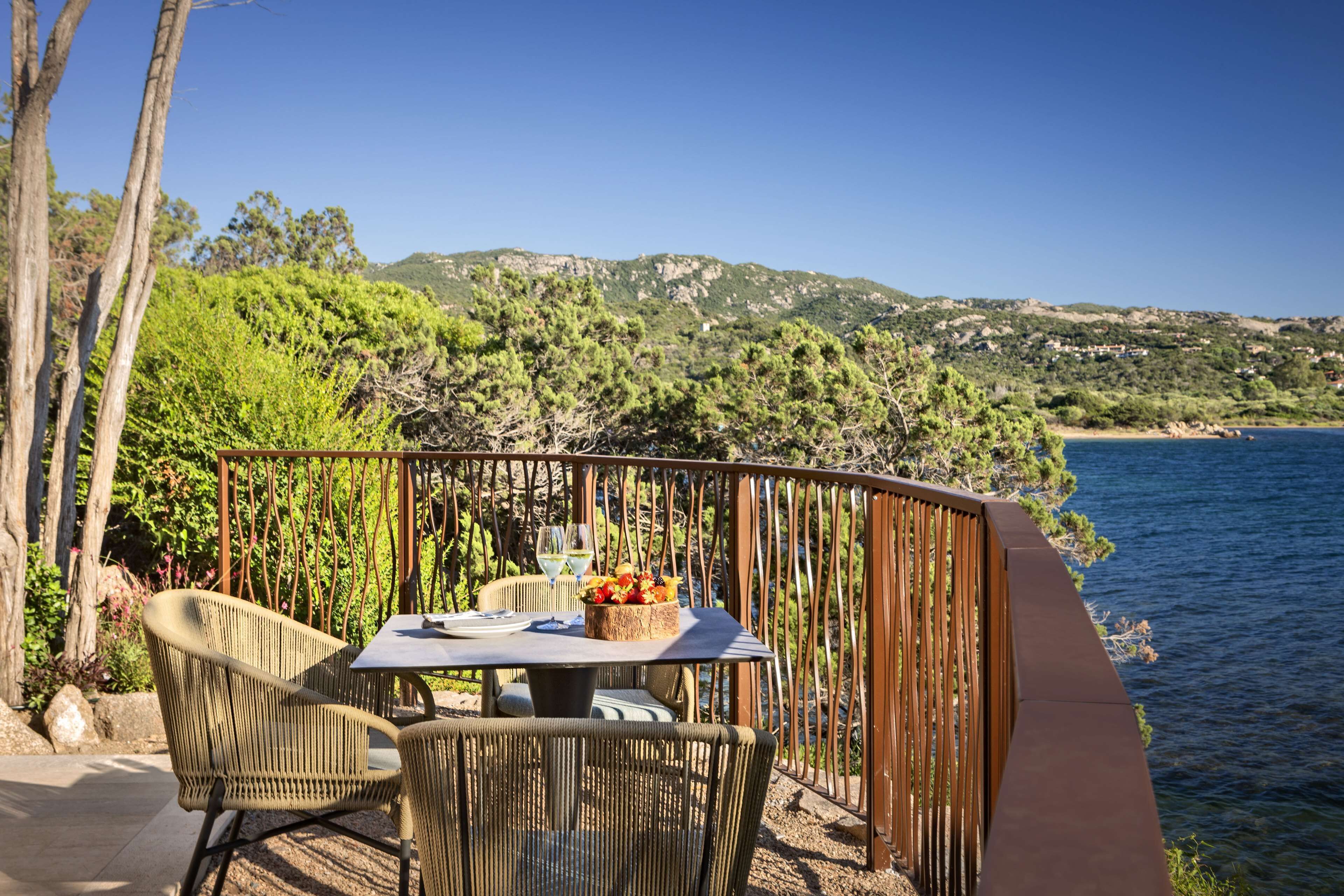 Un espace extérieur aménagé pour les repas, avec des chaises en osier et une table pour deux, offrant une vue sur un paysage côtier composé de collines et d’une étendue d’eau.