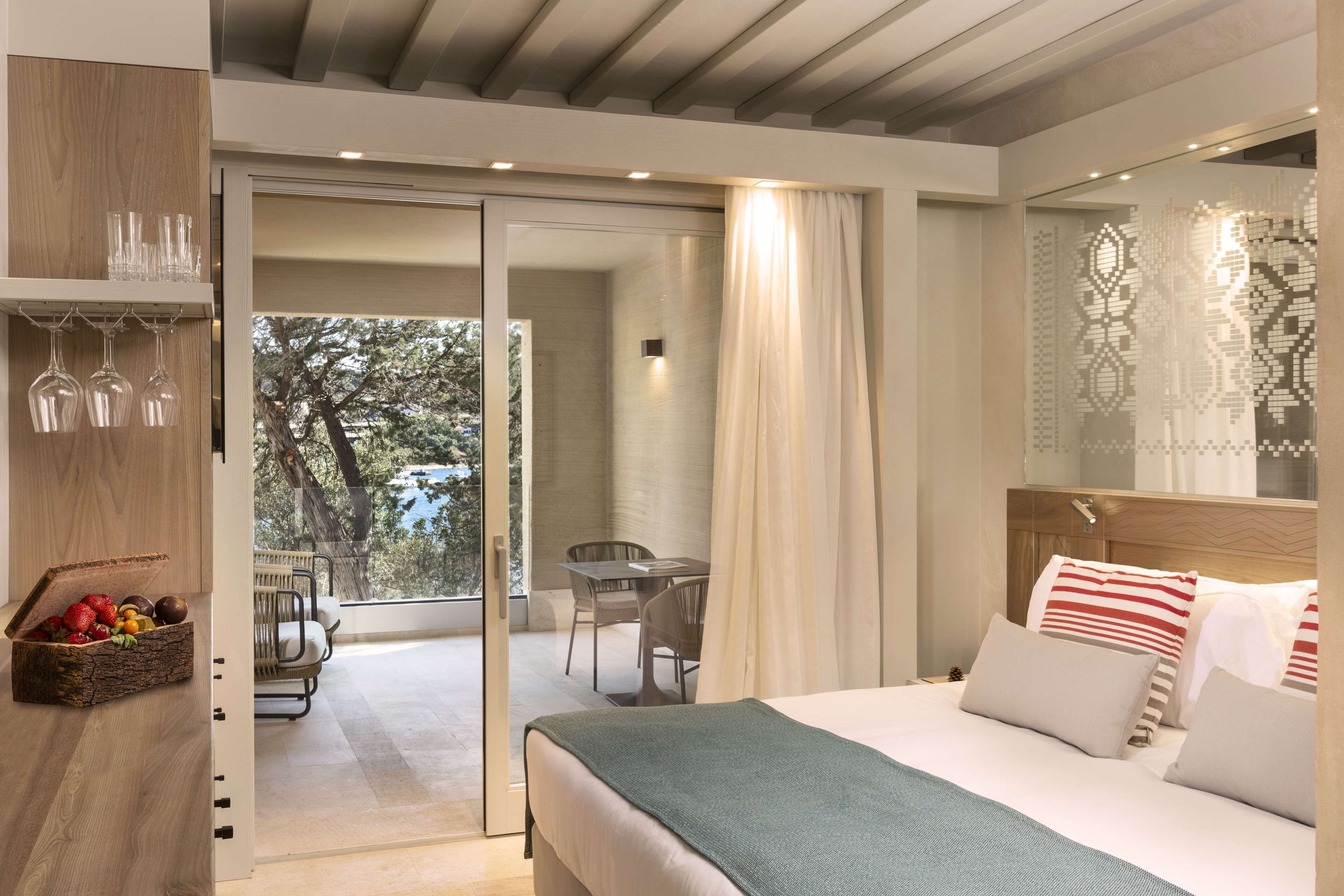 A modern hotel room with a bed, a dining area with a table and chairs, and a balcony with a view of trees.
