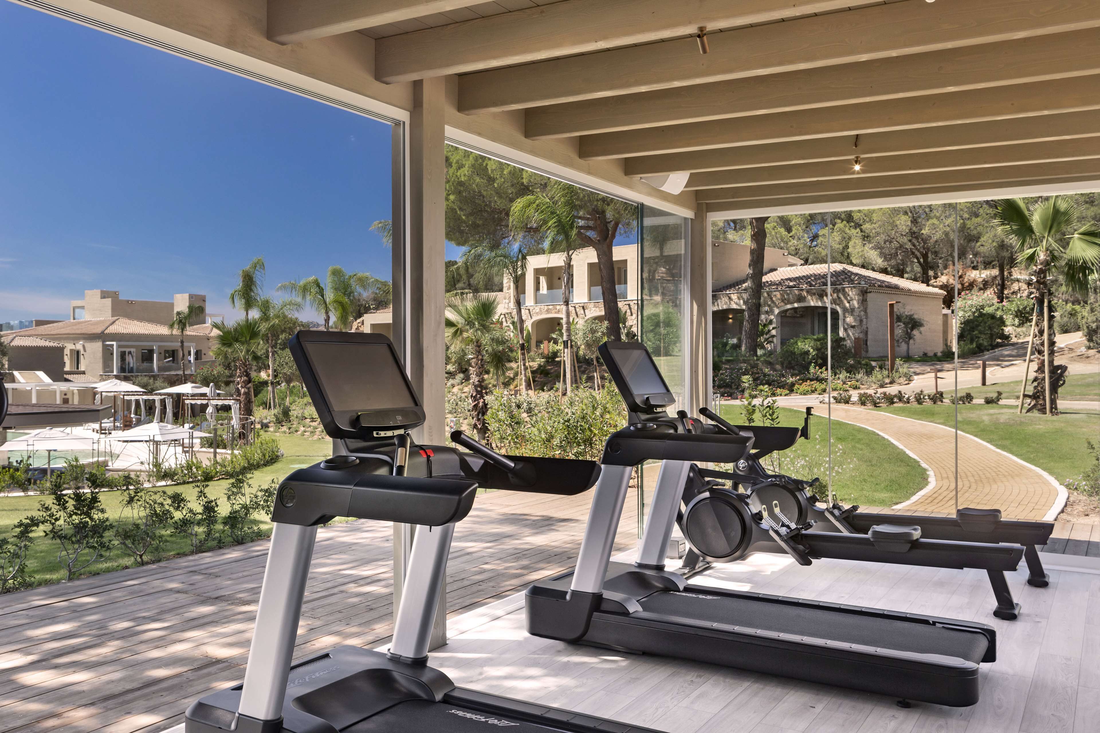 A gym with treadmills and elliptical machines, overlooking a landscaped area with palm trees and a pool.