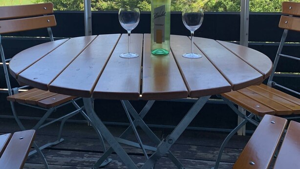 Une table ronde en bois, sur laquelle reposent deux verres à vin et une bouteille de vin Bladrot, est placée sur un balcon offrant une vue sur un paysage verdoyant.
