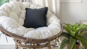Una silla de ratán con un cojín y una almohada oscura, un helecho en maceta y una ventana con cortinas.