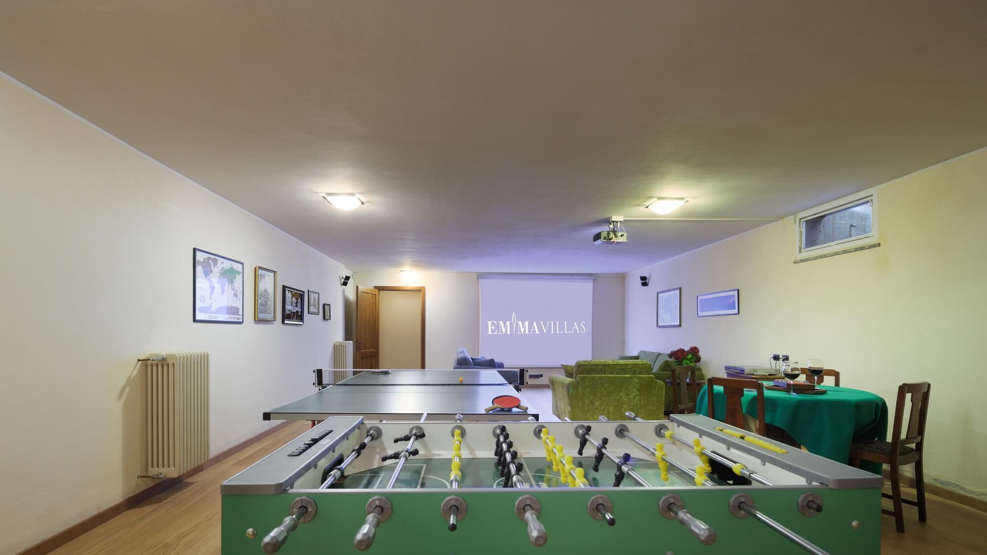 Une pièce avec une table de baby-foot, un écran de projection sur lequel est affiché le texte « EM MAVILLAS », des tableaux encadrés accrochés au mur et une table recouverte d’une nappe verte.