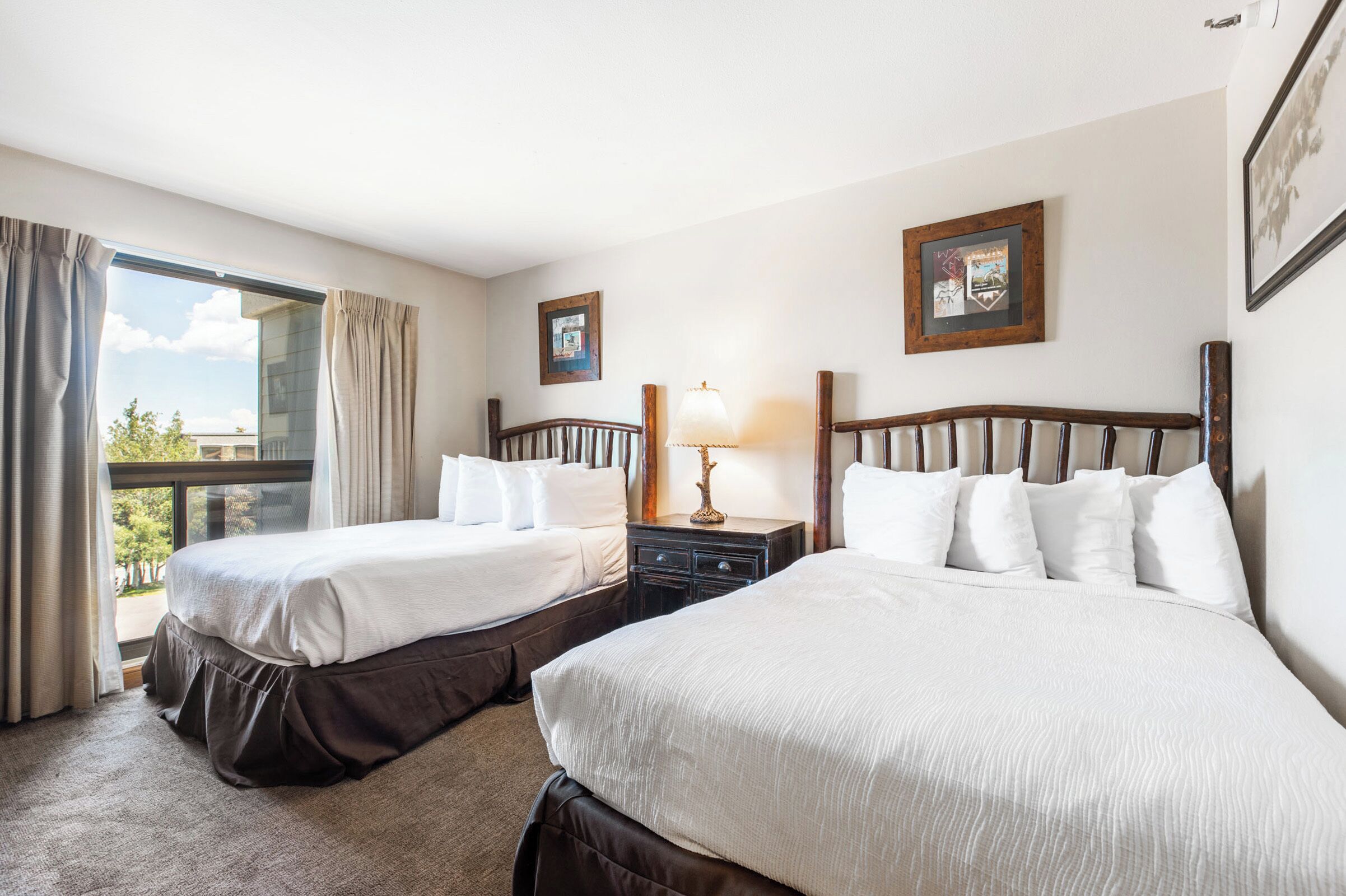 A hotel room with two beds, a nightstand with a lamp, a window with curtains, and framed pictures on the wall.