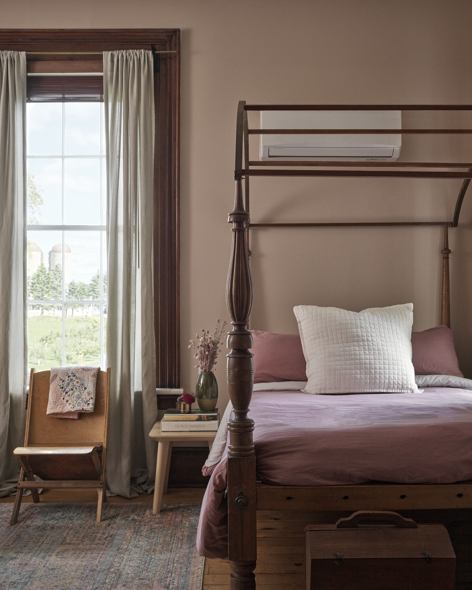 Un dormitorio con una cama con dosel de madera, una mesita de noche con un libro, un jarrón con flores y una ventana con cortinas.