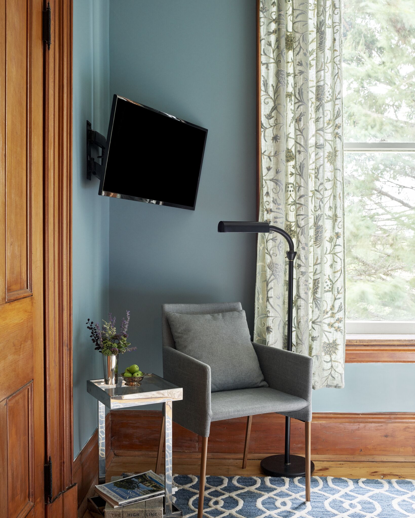 A room with a grey armchair, a small table with a vase of flowers, a mounted flat-screen TV, and a window with patterned curtains.