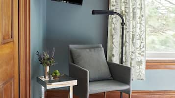 A room with a grey armchair, a small table with a vase of flowers, a mounted flat-screen TV, and a window with patterned curtains.