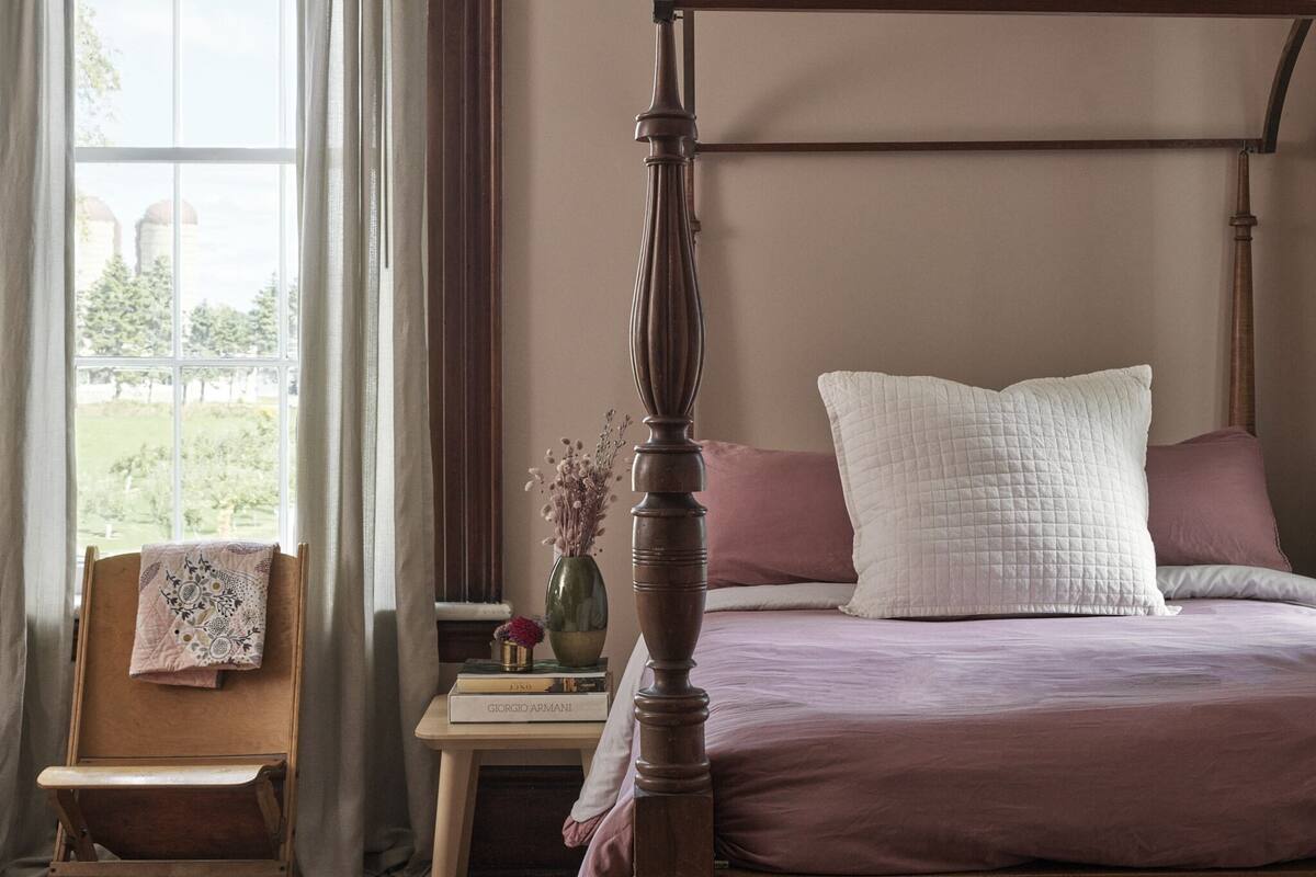 Ein Bett mit Baldachin, ein Holzkopfstück, ein Nachttisch mit Blumenvase und ein Sessel mit gefalteter Decke.