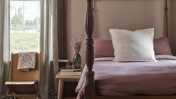 A four-poster bed with a wooden headboard, a bedside table with a vase of flowers, and a chair with a folded blanket.