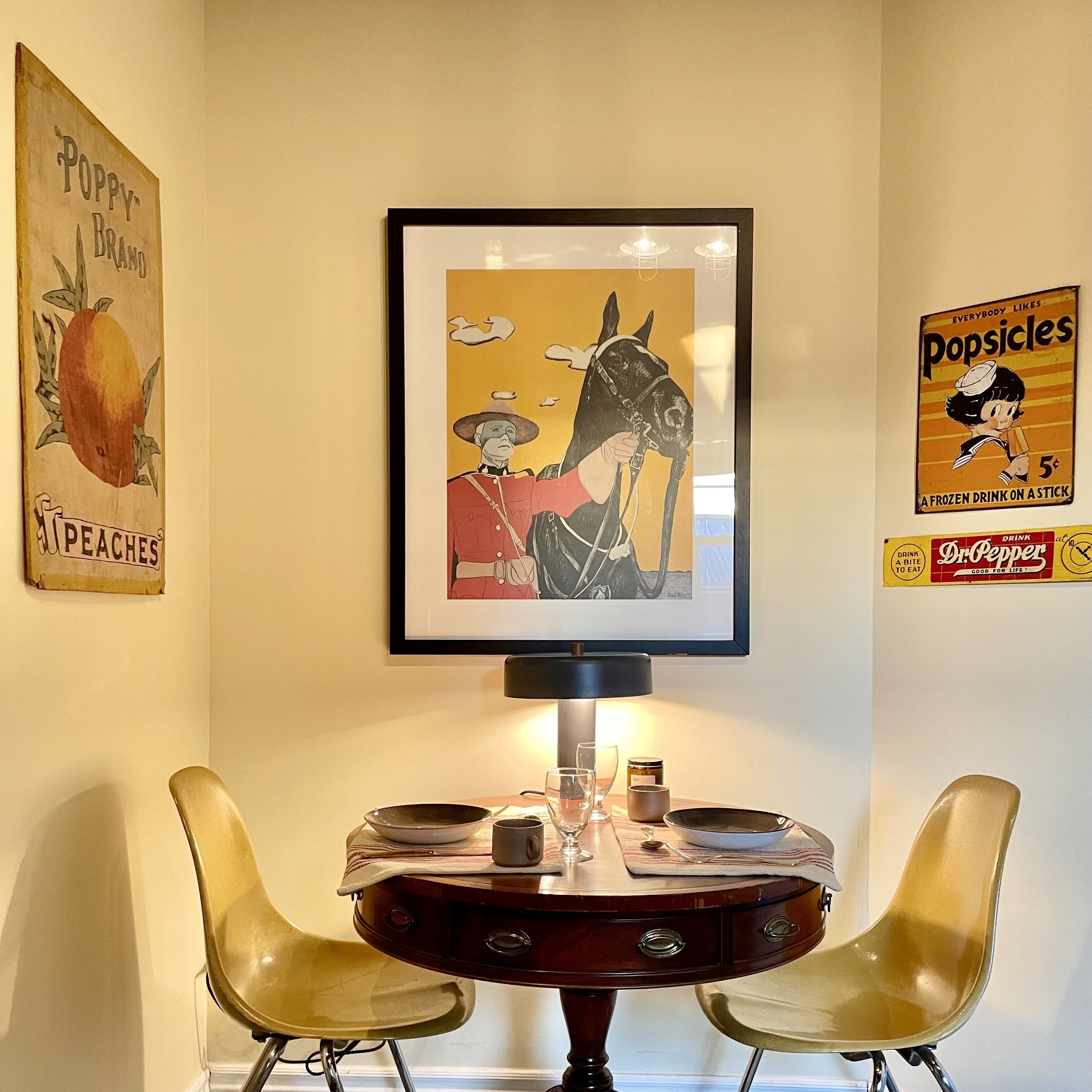 A dining area with a table and two chairs, a framed artwork above the table, and vintage posters on the wall.