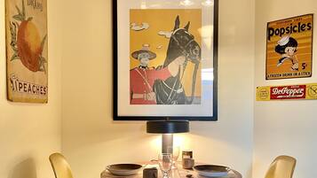 A dining area with a table and two chairs, a framed artwork above the table, and vintage posters on the wall.
