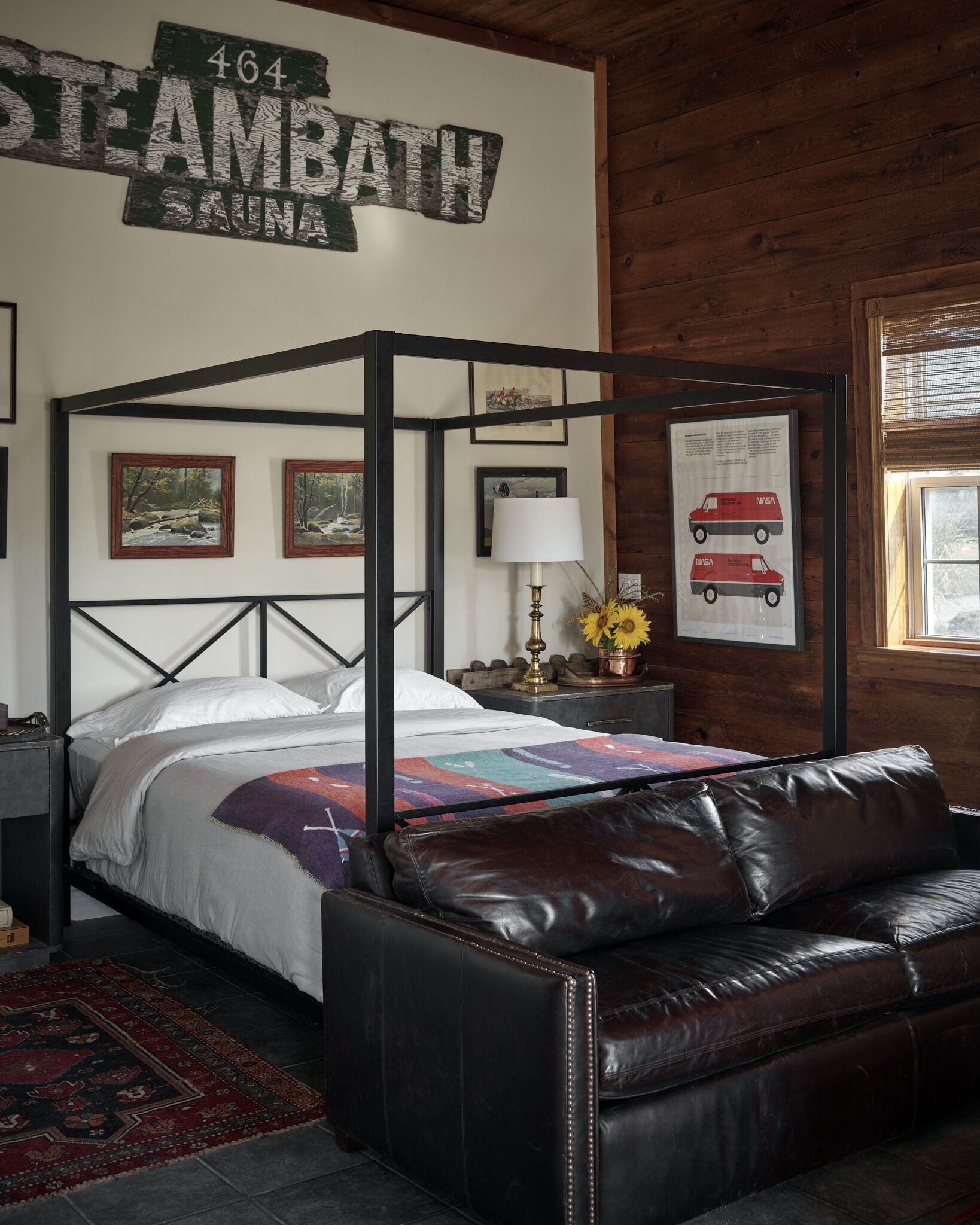 A four-poster bed with a colorful quilt, a leather sofa, a wooden wall with a rustic sign, framed pictures, and a lamp with sunflowers.