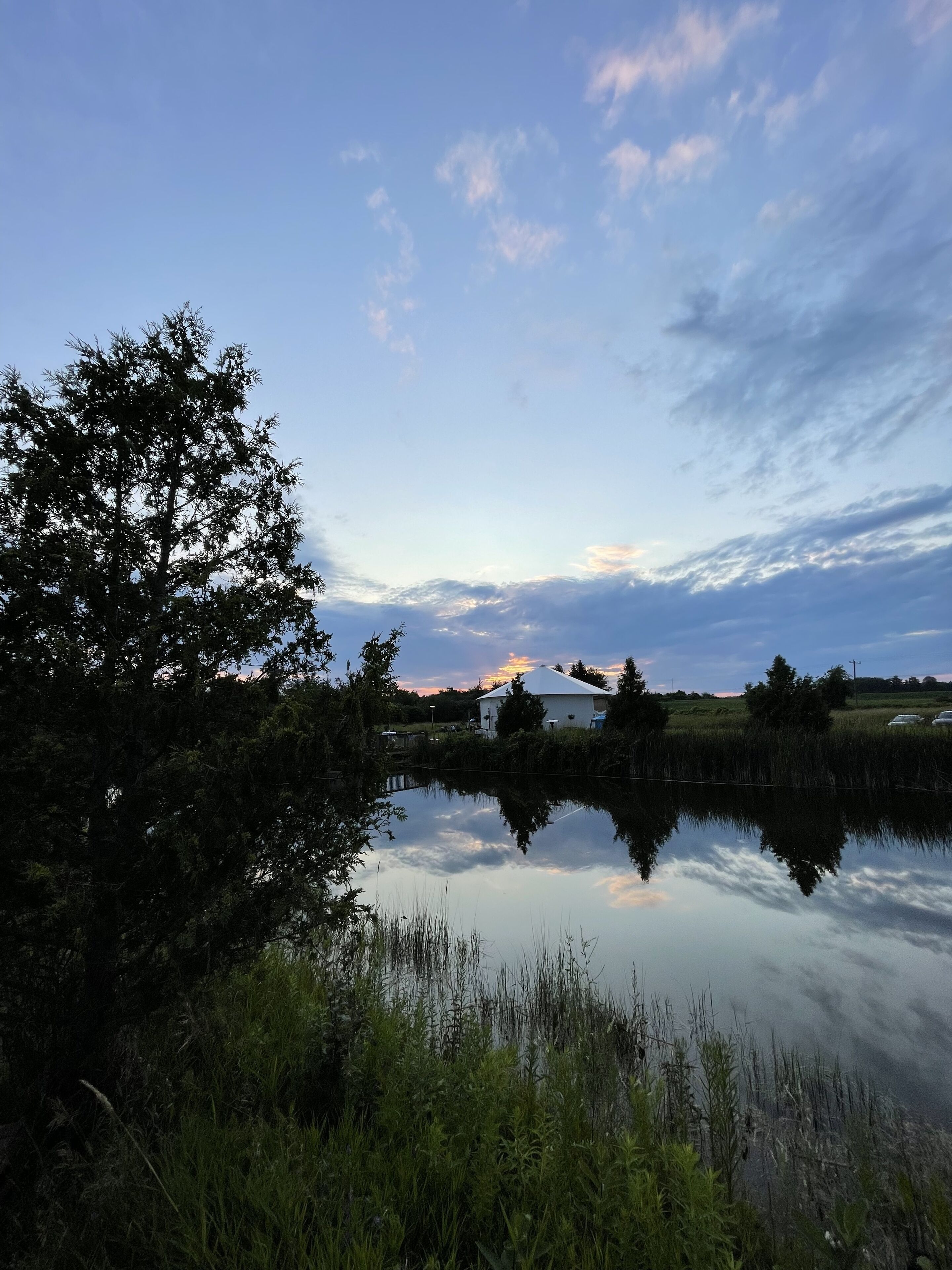 A tranquil lakeside scene with a house in the background, surrounded by lush greenery and a clear sky.