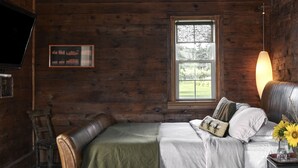 A wooden cabin bedroom with a bed, a window, a lamp, and a TV mounted on the wall. - The Eddie Hotel and Farm (Prince Edward)