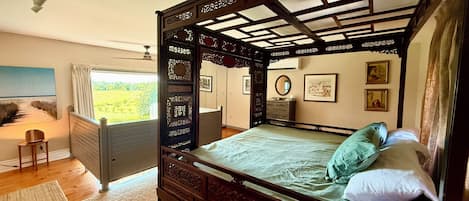 A four-poster bed with a wooden frame and intricate carvings, a small bedside table, a radiator, and a painting on the wall.