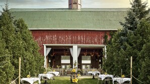 An outdoor wedding venue with a rustic barn, multiple tables with white tablecloths, and a central decorative barrel. - The Eddie Hotel and Farm (Prince Edward)