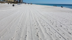 Une vaste plage de sable fin, parsemée d’empreintes qui mènent vers l’océan, avec des bâtiments en arrière-plan et un ciel bleu dégagé.