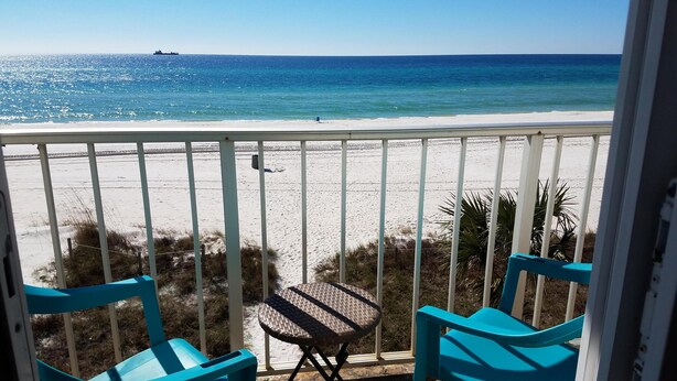 Un balcon avec des chaises turquoise et une petite table, donnant sur une plage où l’on aperçoit un navire au loin.