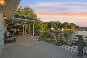 A covered deck with outdoor seating, a view of a lake, and a distant shoreline with buildings.