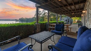 Een overdekt terras met tuinmeubilair, een tafel en uitzicht op een meer bij zonsondergang.