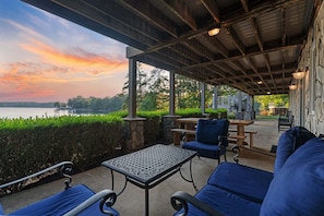Eine überdachte Terrasse mit Gartenmöbeln, einem Tisch und Blick auf einen See bei Sonnenuntergang.